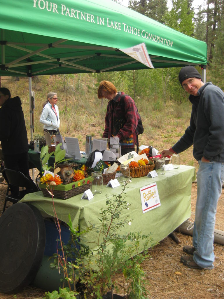 Image shows a green canopy tent with the words "Your partner in Lake Tahoe Conservation". Several people are surrounding the canopy looking at outreach materials that are on a table underneath the canopy tent.