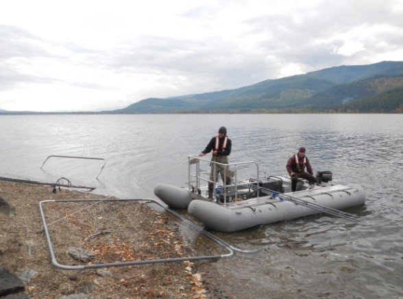 Image shows two people in an inflatebale boat at a lake's shore. The boat has a large square benthic electrode array that the men are preparing for deployment.