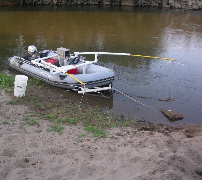 Image shows a boat with an electrofishing apparatus sttached to it. The apparatus has wires that dangle into the water and is connected to the boat with white pvc pipe.