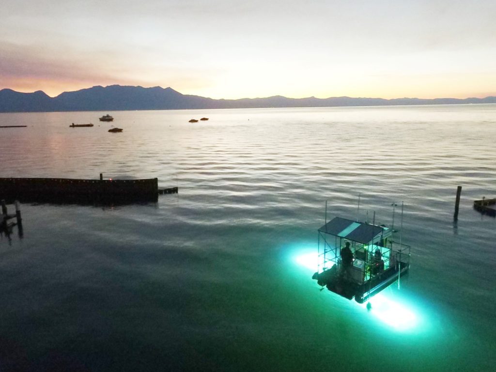 Image shows overhead show of Lake Tahoe at sunset. A boat is in the foreground with green UV light shining into the water below it. 